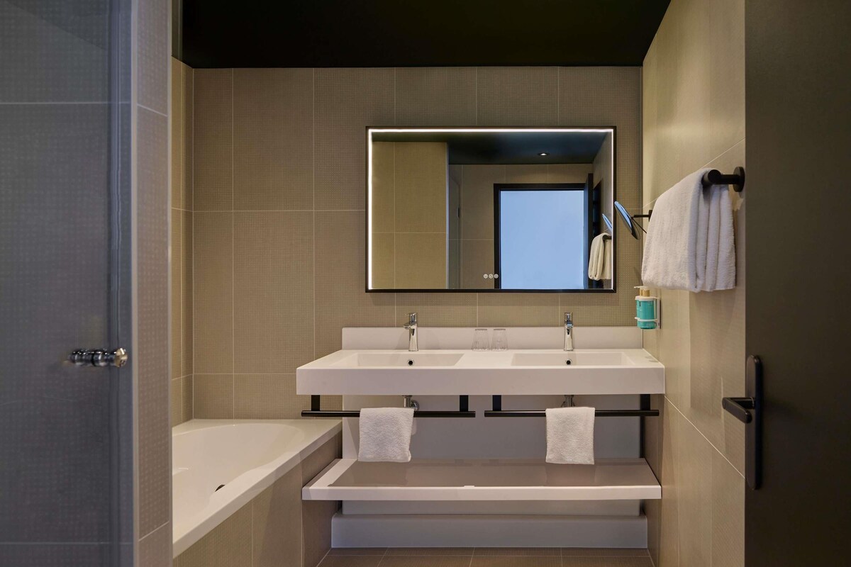 A modern bathroom with a large mirror, two sinks, a bathtub, and a towel rack.