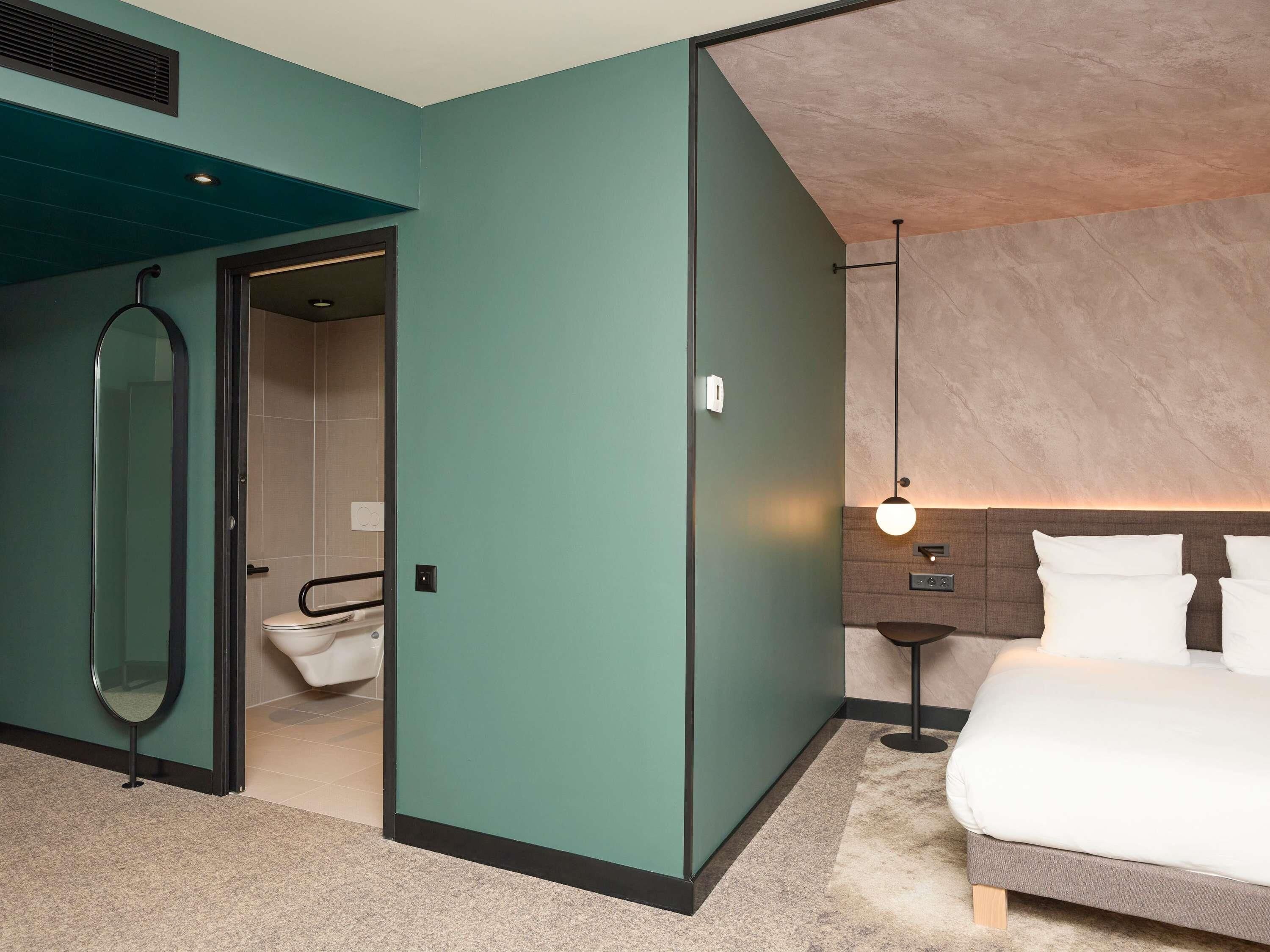 Quarto de hotel com cama, mesa de cabeceira, luminária na parede, espelho e porta para o banheiro.