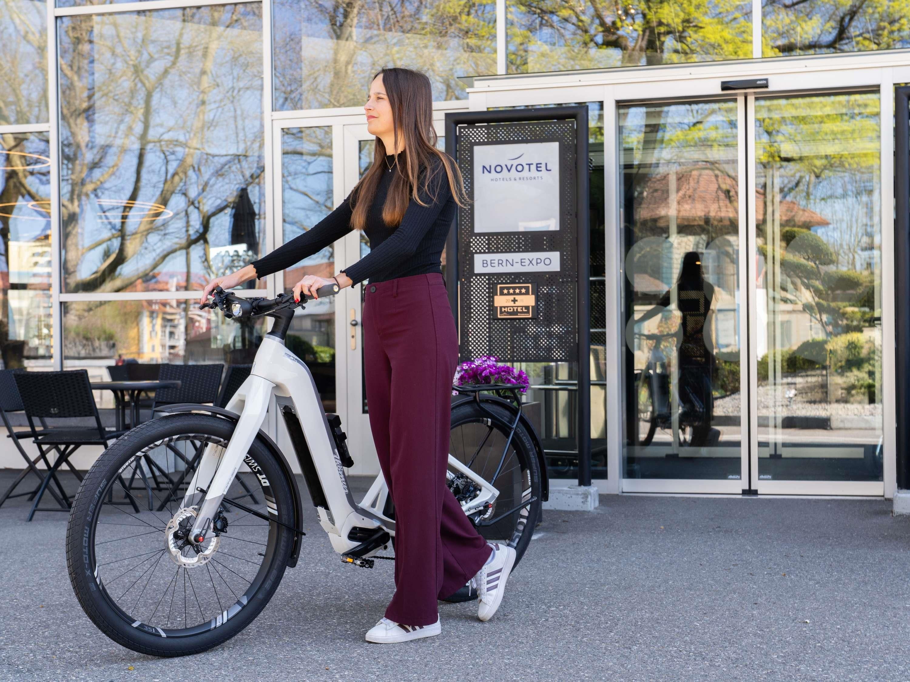 Une personne est debout devant l'entrée d'un hôtel, à côté d'un vélo électrique blanc. On peut voir une enseigne indiquant « Novotel Bern-Expo ».