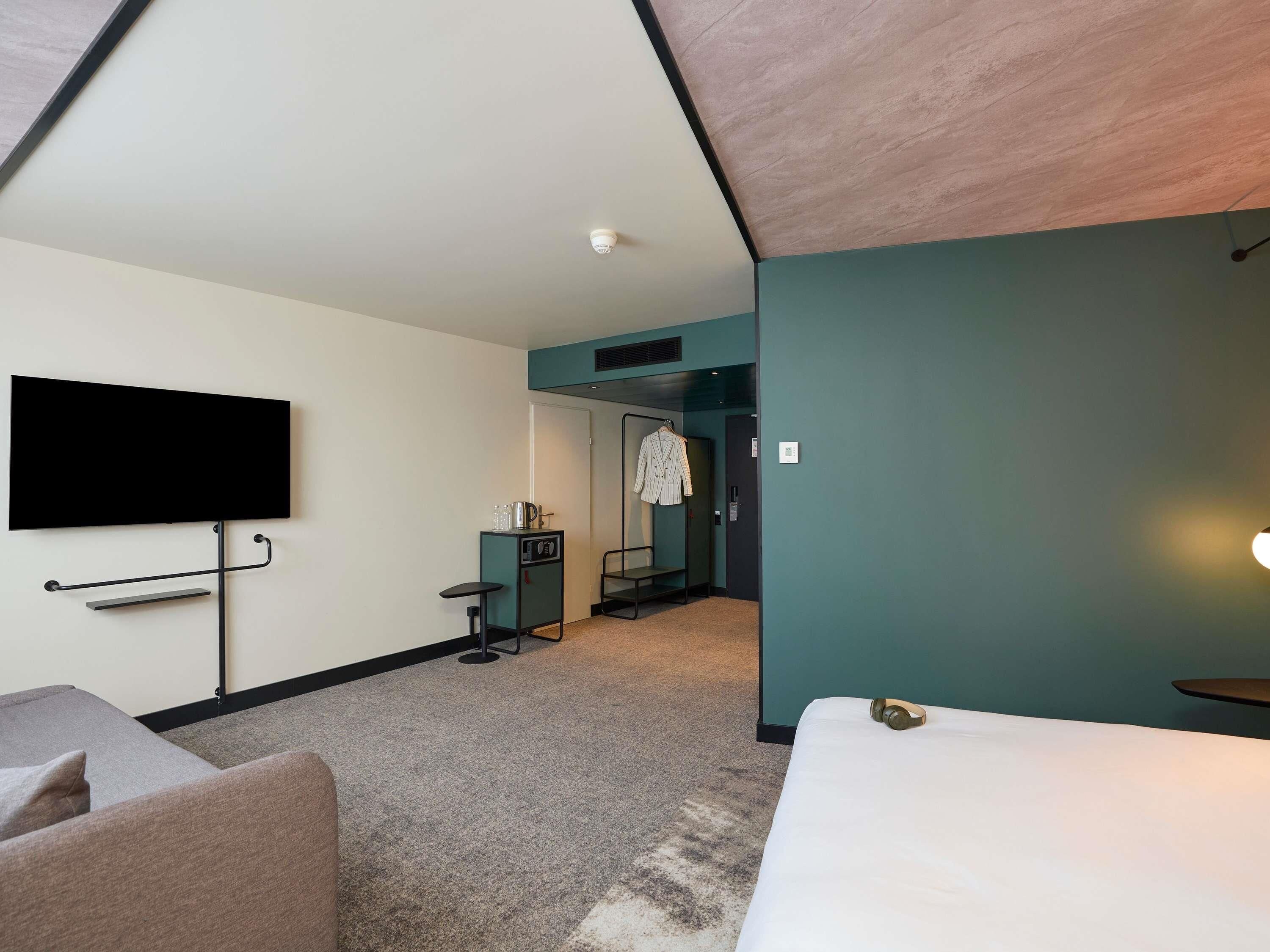 Une chambre d’hôtel moderne équipée d’une télévision à écran plat, d’un lit, d’un petit bureau avec une chaise et d’un porte-manteau.