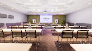 Un salón de conferencias con una gran pantalla mostrando 'NOVOTEL' y 'BERN EXPO' al frente. Hay varias mesas y sillas dispuestas para reuniones.