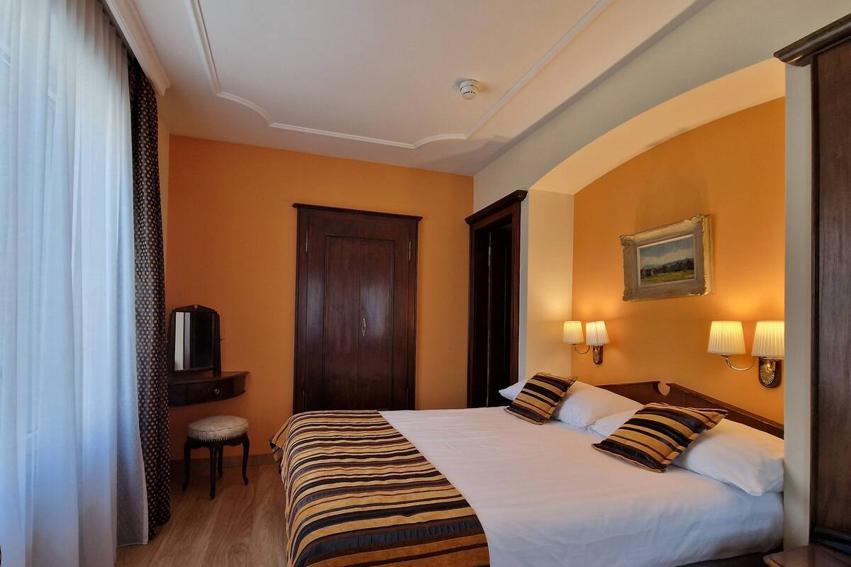 Habitación de hotel con una cama grande, dos lámparas de noche, un armario de madera, una mesita con una silla y un televisor sobre un mueble.