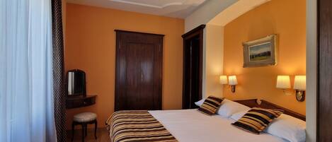 Habitación de hotel con una cama grande, dos lámparas de noche, un armario de madera, una mesita con una silla y un televisor sobre un mueble.