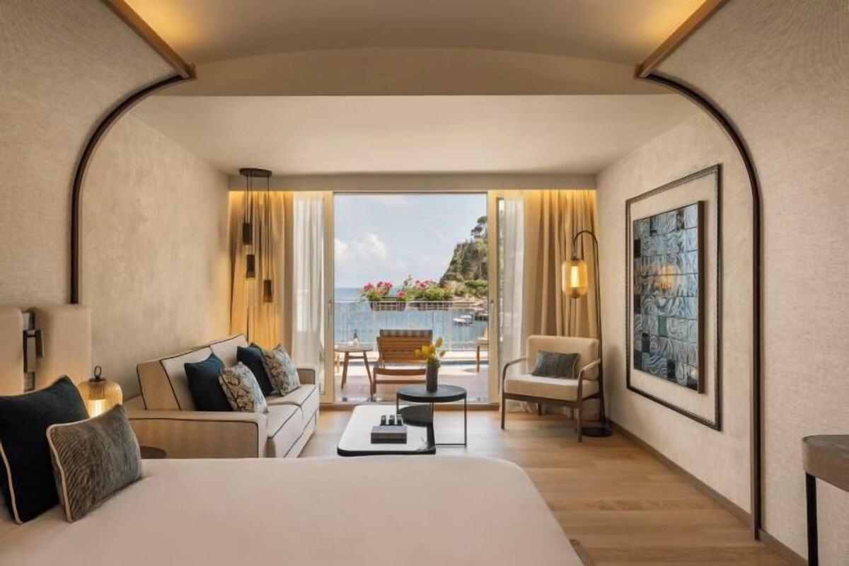 Een hotelkamer met een groot bed, een bank, een klein tafeltje en een balkon met uitzicht op zee.