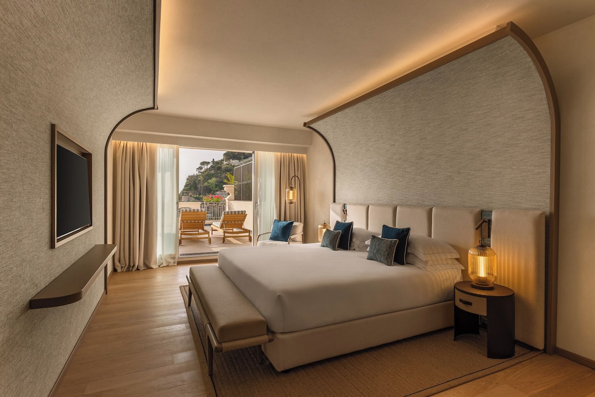 Une chambre d’hôtel moderne avec un grand lit, un téléviseur fixé au mur, une table de chevet avec une lampe et une vue sur l’extérieur grâce à une porte coulissante.