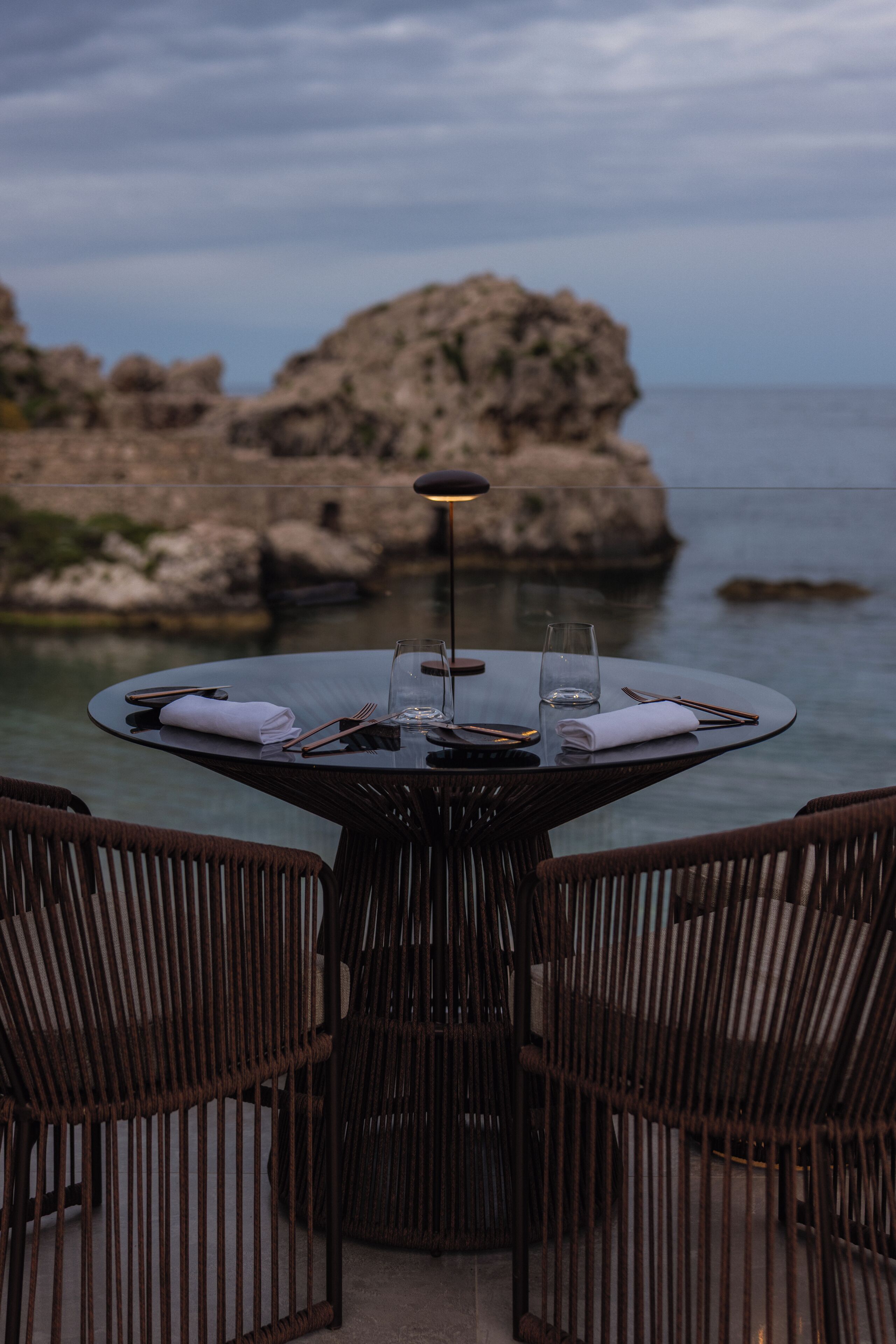 A round table with a glass top, surrounded by wicker chairs, set against a rocky coastal backdrop.