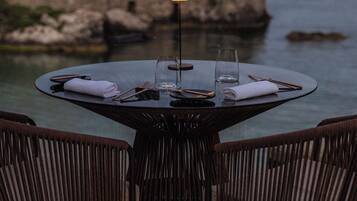 A round table with a glass top, surrounded by wicker chairs, set against a rocky coastal backdrop.