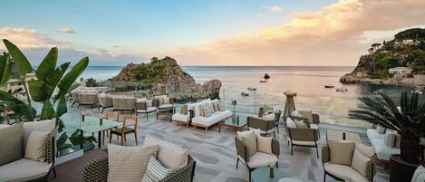 A terrace with outdoor seating, overlooking a coastal area with boats and rocky terrain.
