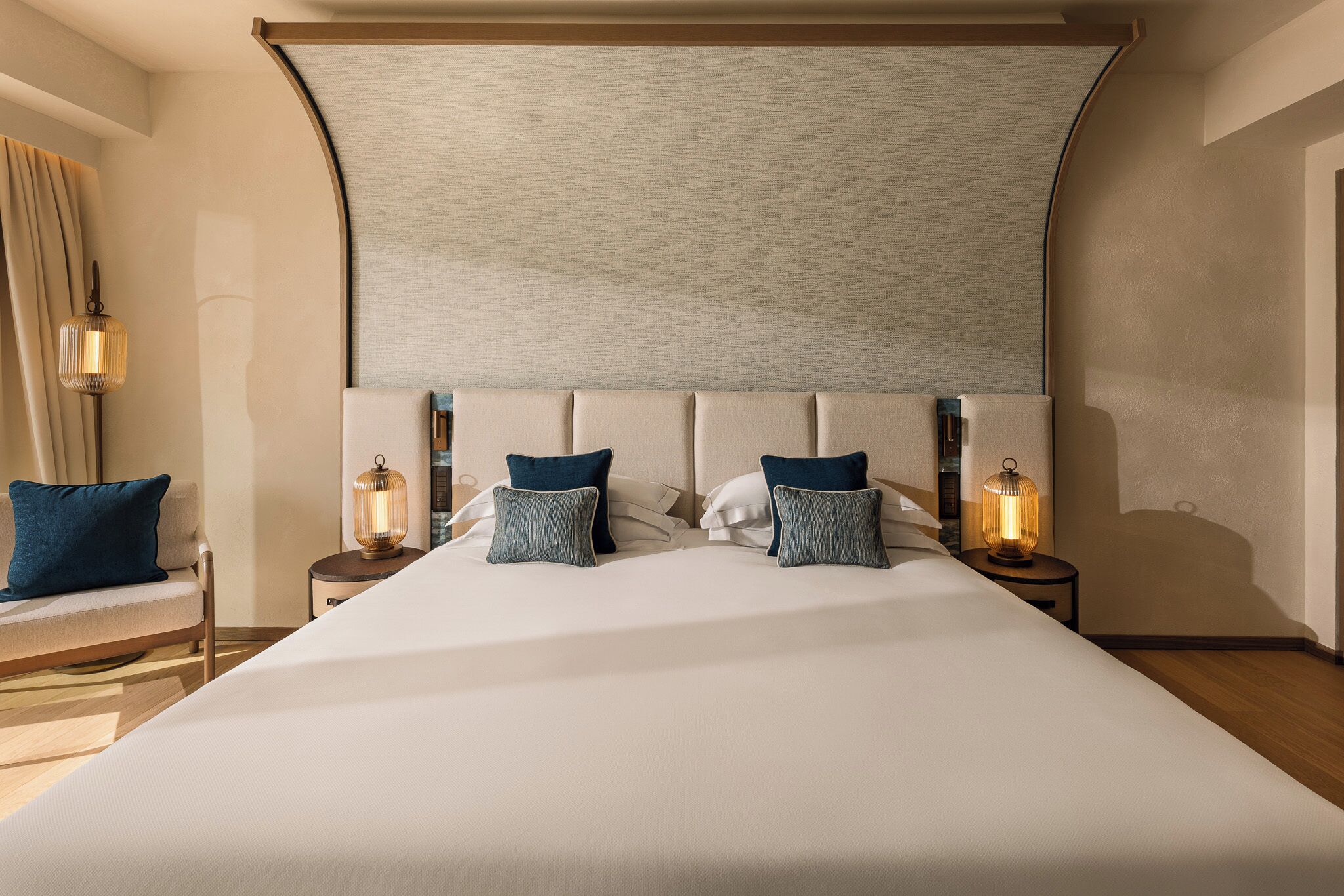Een opgemaakt bed met hoofdeinde, twee nachtkastjes met lampen, en een stoel met kussen.