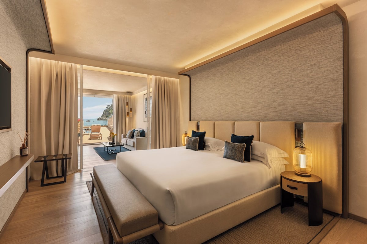 Une chambre d’hôtel avec un grand lit, des tables de chevet, un banc, une vue sur la mer et un balcon.