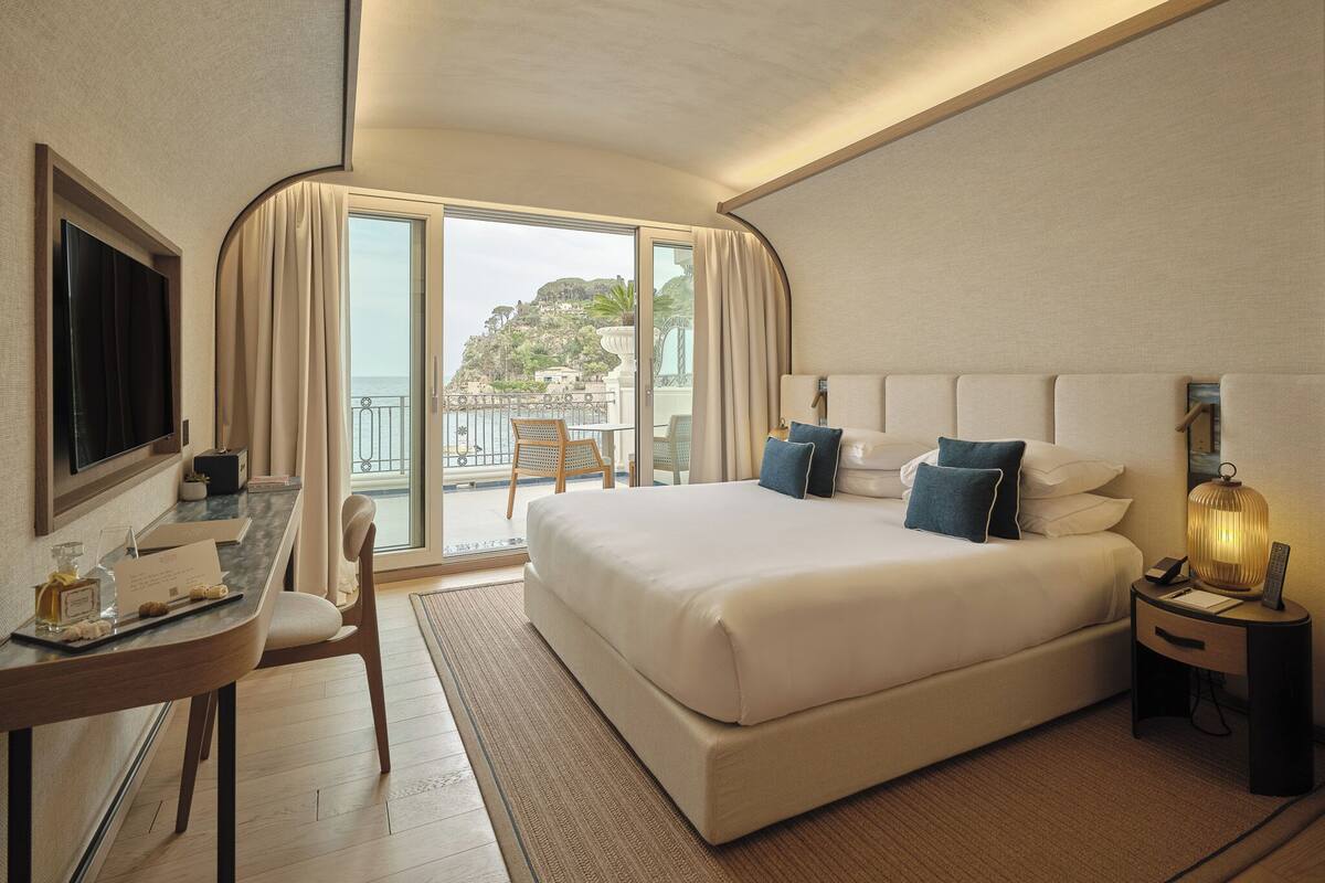 Une chambre d’hôtel comprenant un lit, un bureau, une chaise, une télévision, une table de chevet et un balcon avec vue sur une colline.