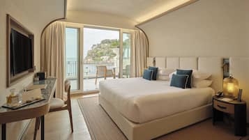 A hotel room with a bed, a desk, a chair, a TV, a nightstand, and a balcony with a view of a hill.
