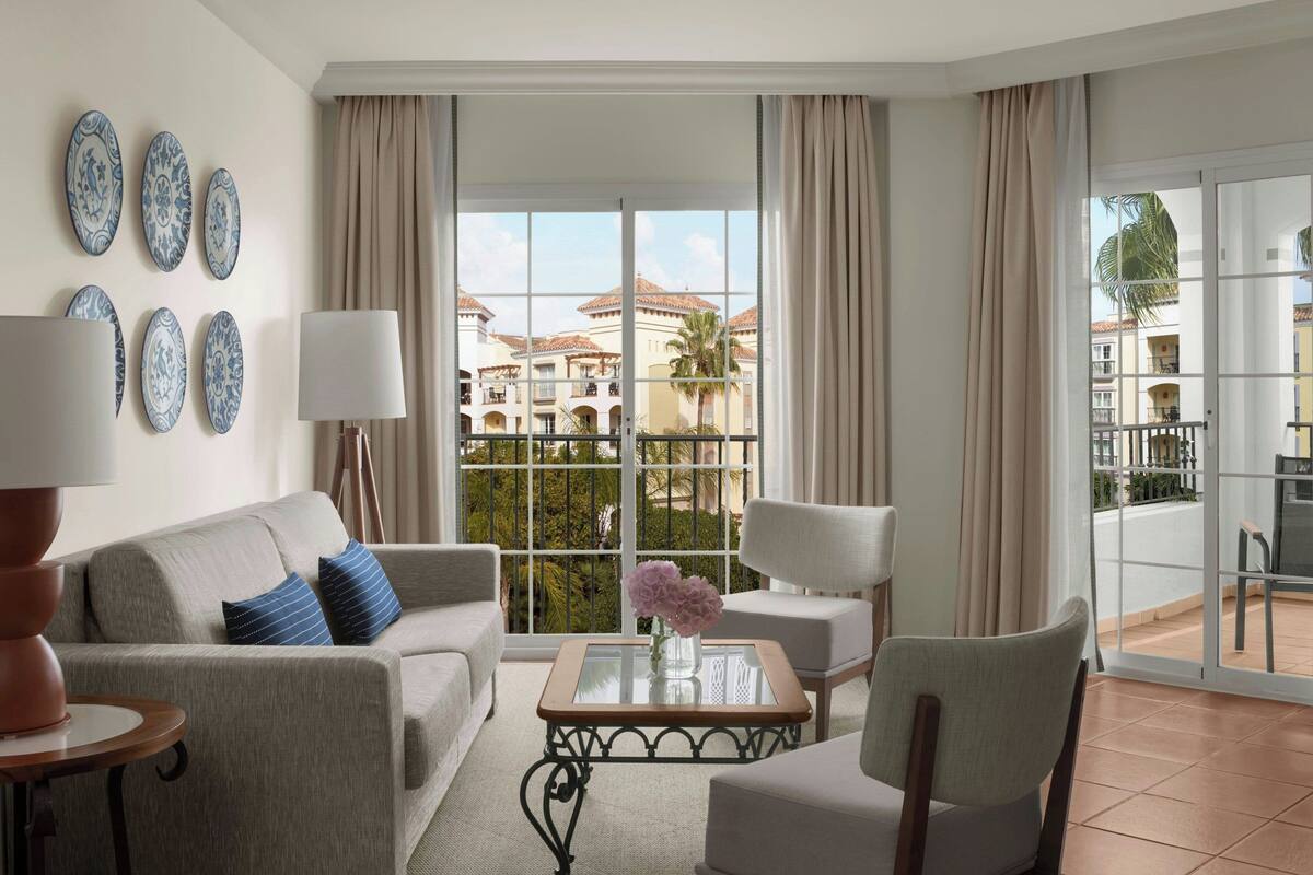 A hotel room with a balcony, a sofa, a chair, a coffee table, and decorative plates on the wall.