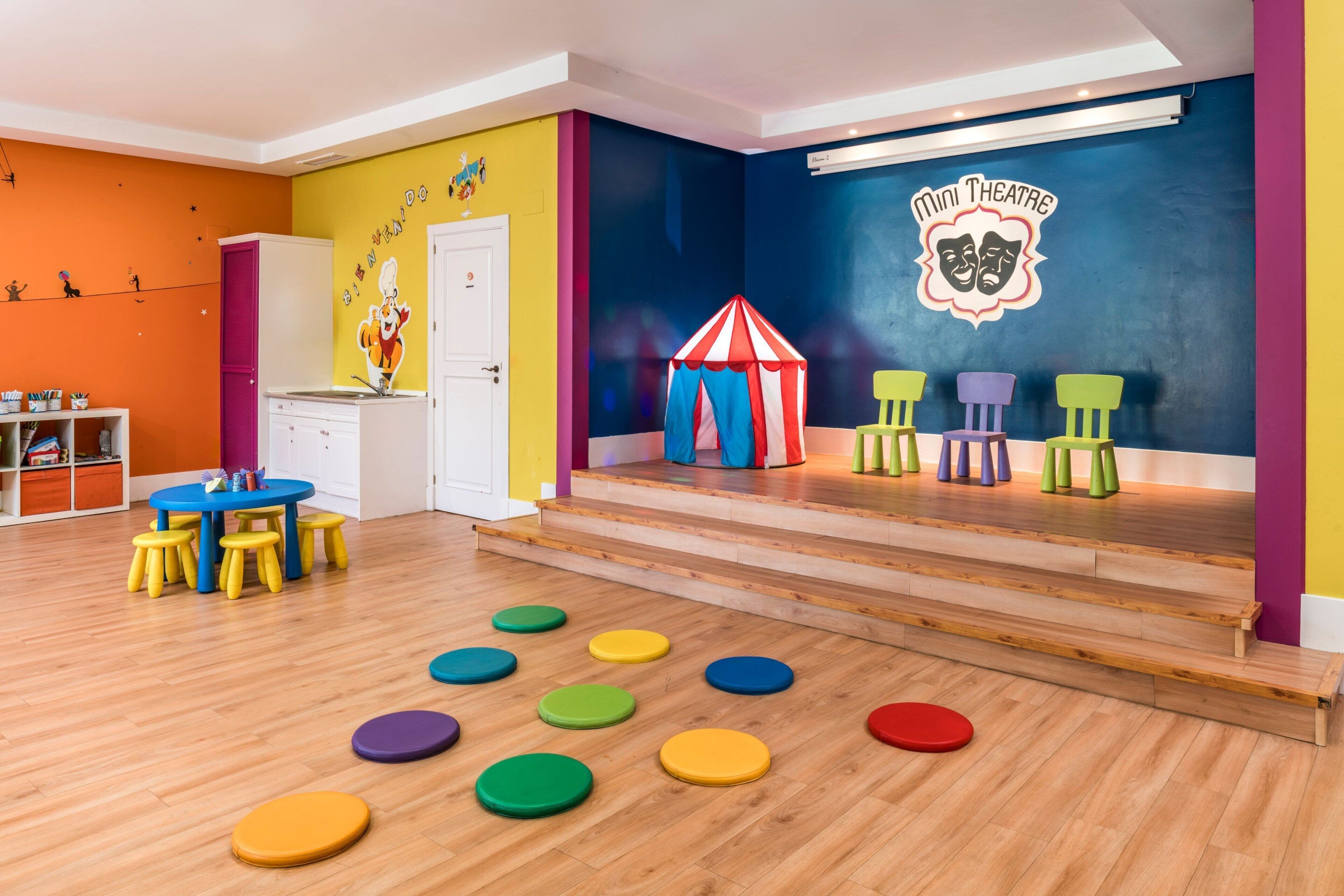 A colorful playroom with a mini theatre, wooden flooring, and a variety of stools and tables.
