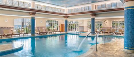 An indoor swimming pool with a clear view of the sky through the ceiling, surrounded by lounge chairs and a tiled floor.