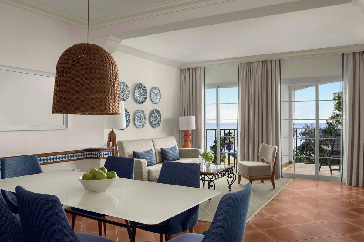 A dining area with a white table, blue chairs, a wicker pendant light, and a bowl of fruit.