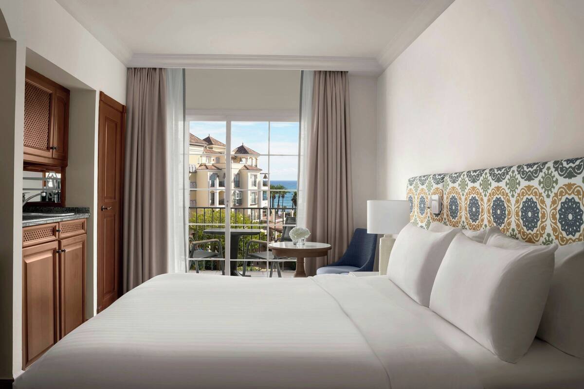 A hotel room with a large bed, a dining table, a chair, a view of the sea, and a balcony.