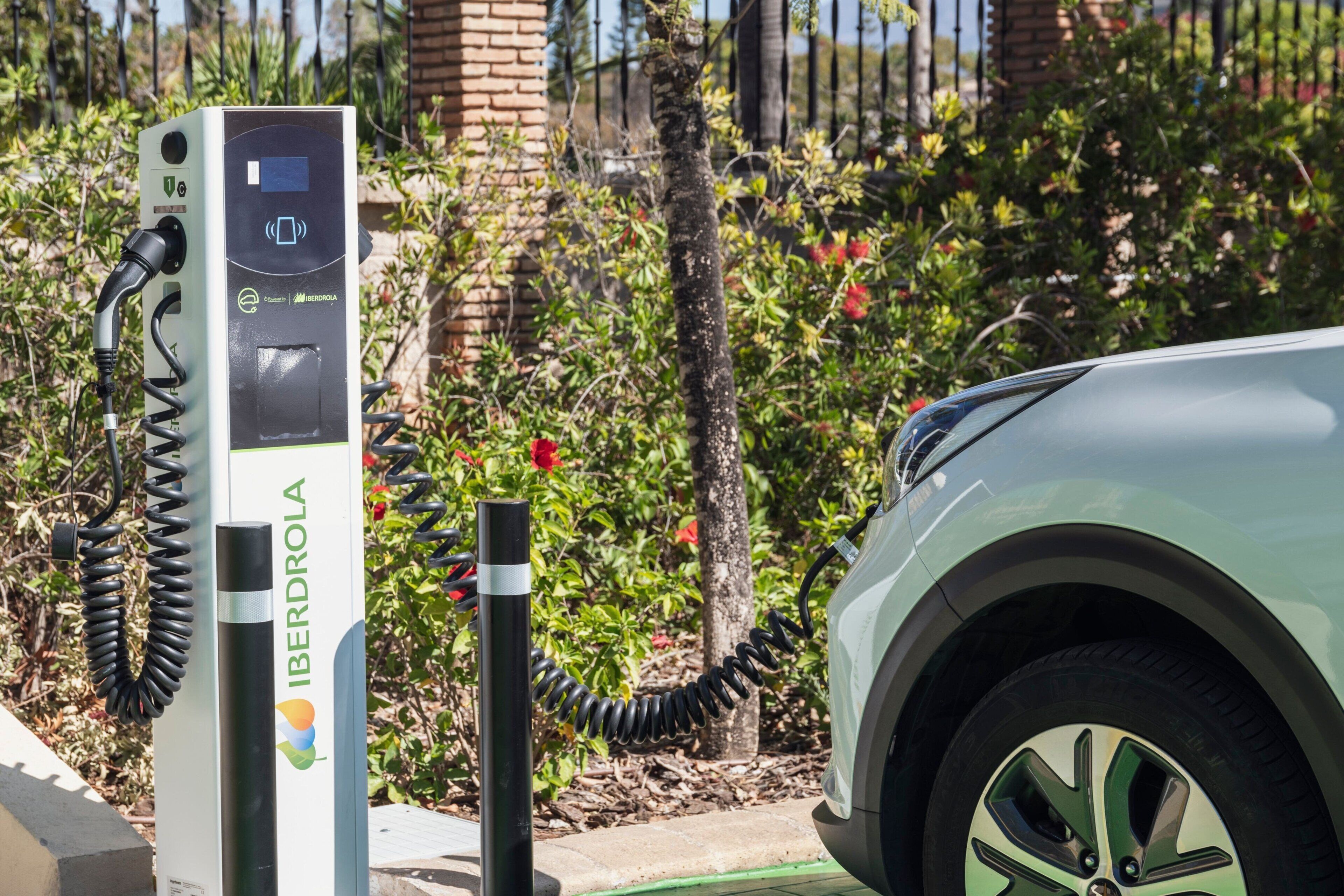 Una estación de carga para vehículos eléctricos, con un automóvil conectado, rodeada de follaje y una cerca.
