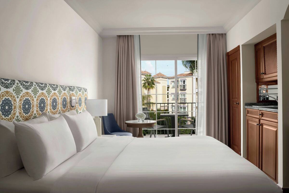 A hotel room with a bed, a chair, a small table, a balcony with a view of buildings, and wooden cabinets.