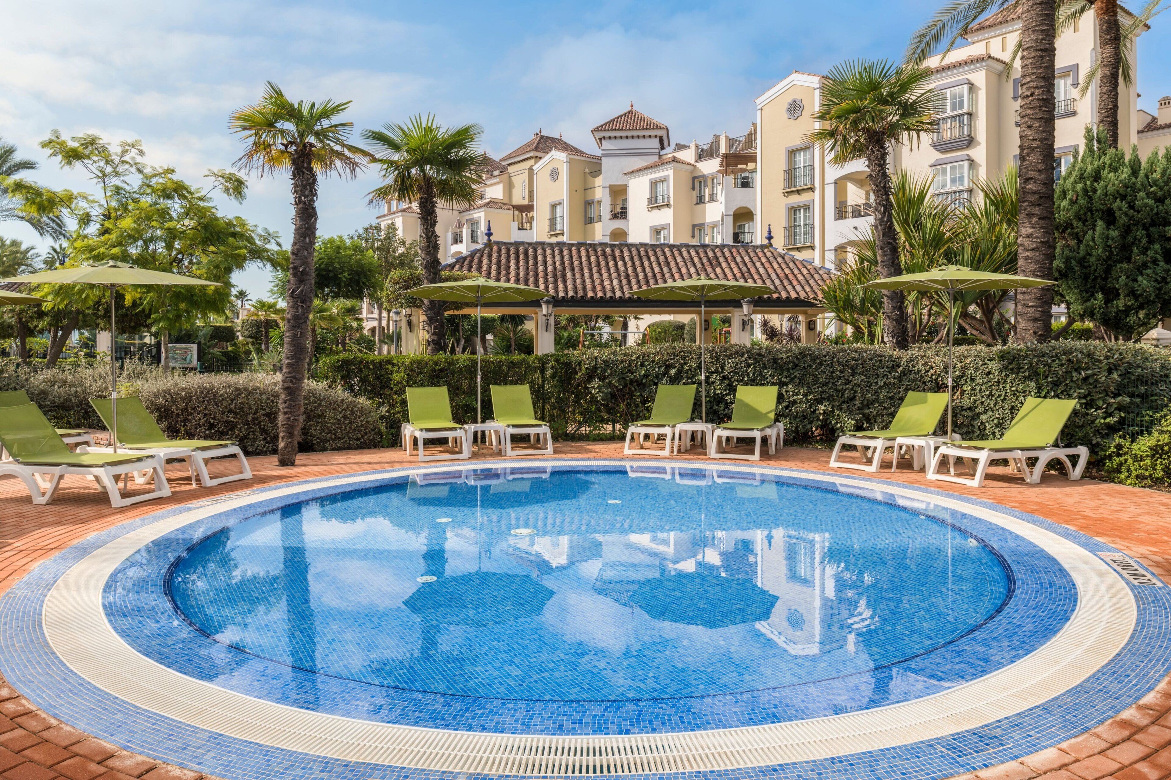 A pool surrounded by lounge chairs and umbrellas, with a building in the background.