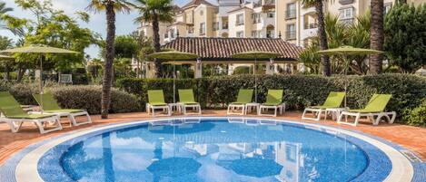 A pool surrounded by lounge chairs and umbrellas, with a building in the background.