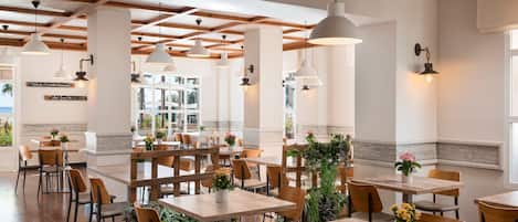 A spacious dining area with wooden tables and chairs, pendant lighting, and large windows offering a view of the outdoors.