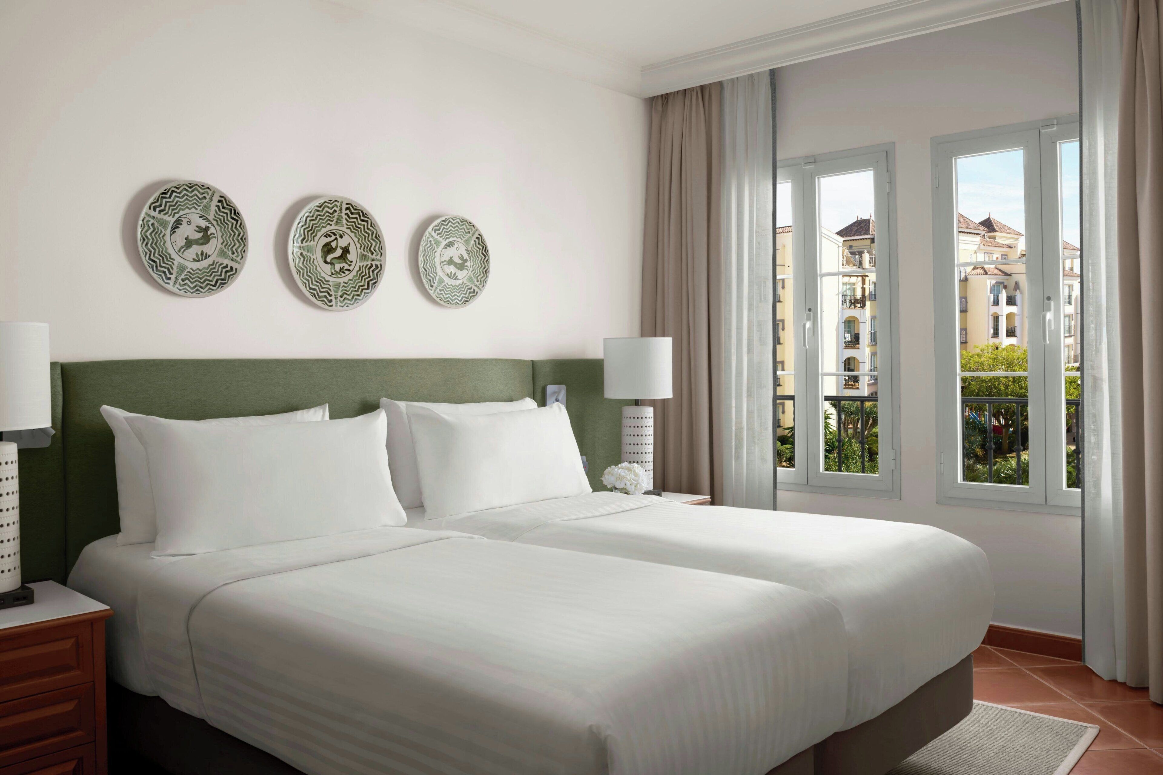 Ein Hotelzimmer mit zwei Betten, einem grünen Kopfteil, Wanddekorationen und Blick auf Gebäude durch große Fenster.
