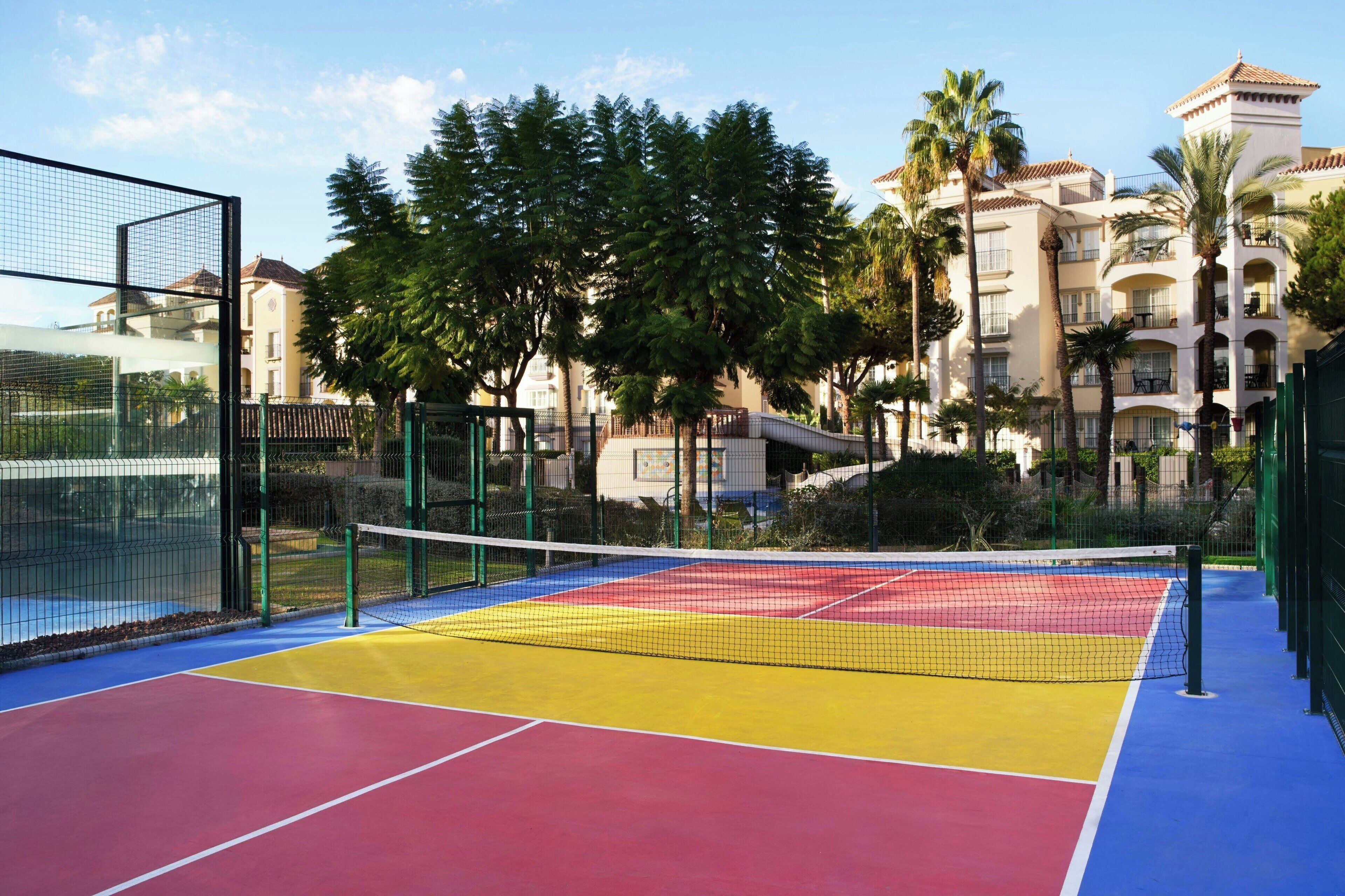 Een tennisbaan met een net, omringd door een hek, en gebouwen op de achtergrond.