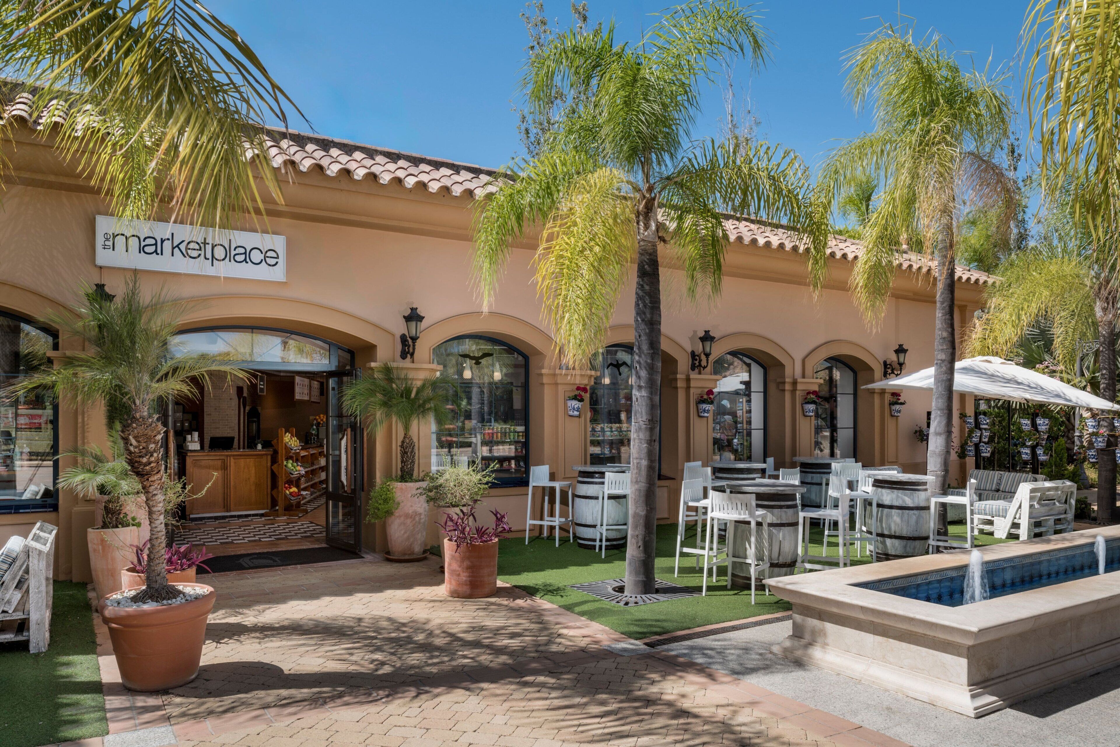 Une devanture de magasin avec une enseigne indiquant « The Marketplace », des tables et des chaises à l’extérieur, protégées par des parasols, et une petite piscine.