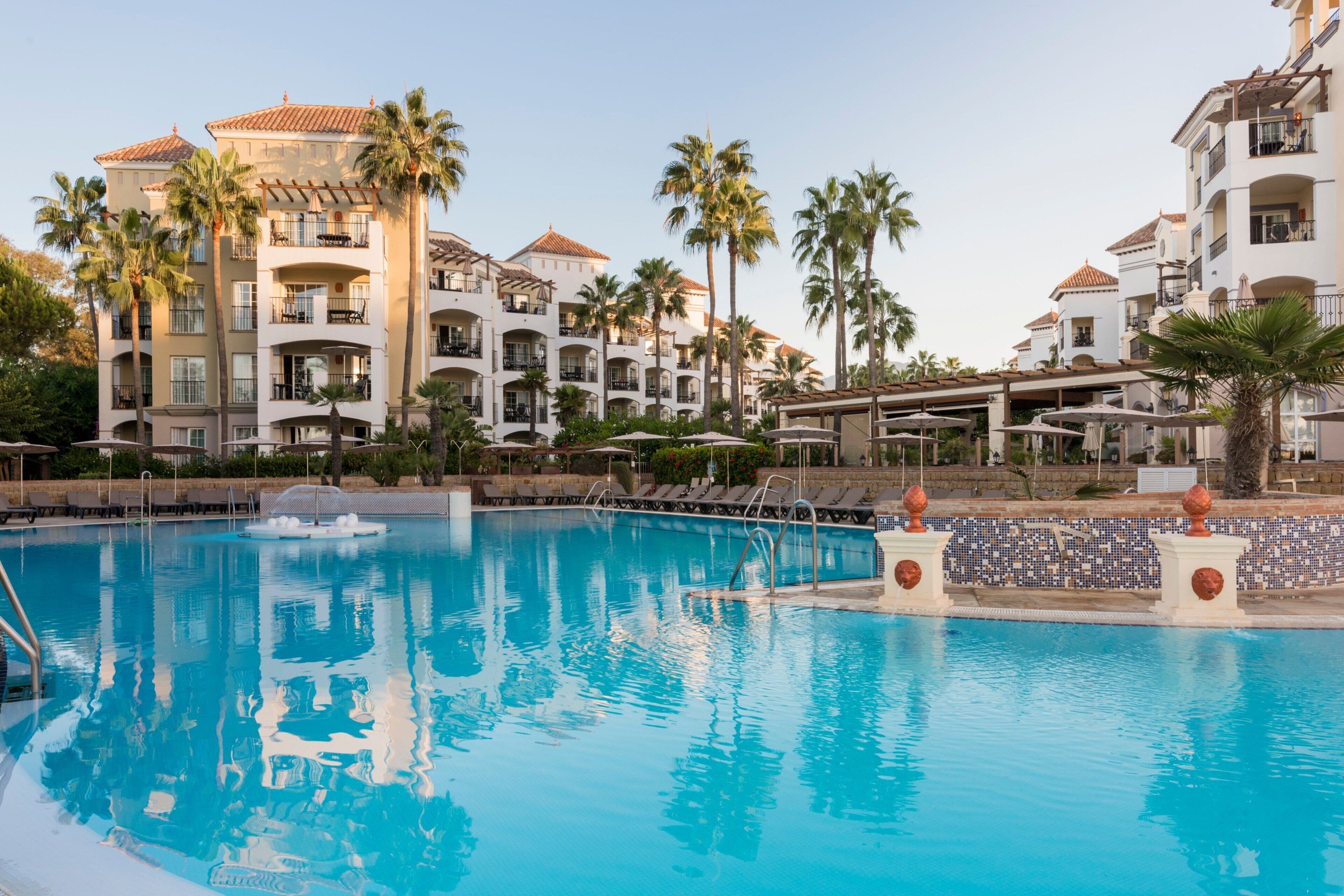 A swimming pool with lounge chairs and umbrellas, surrounded by multi-story buildings with balconies and palm trees.