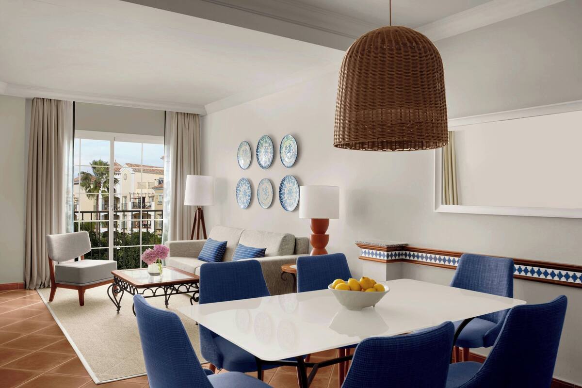 A hotel room with a dining area, a sofa, a coffee table, and a view of the city.