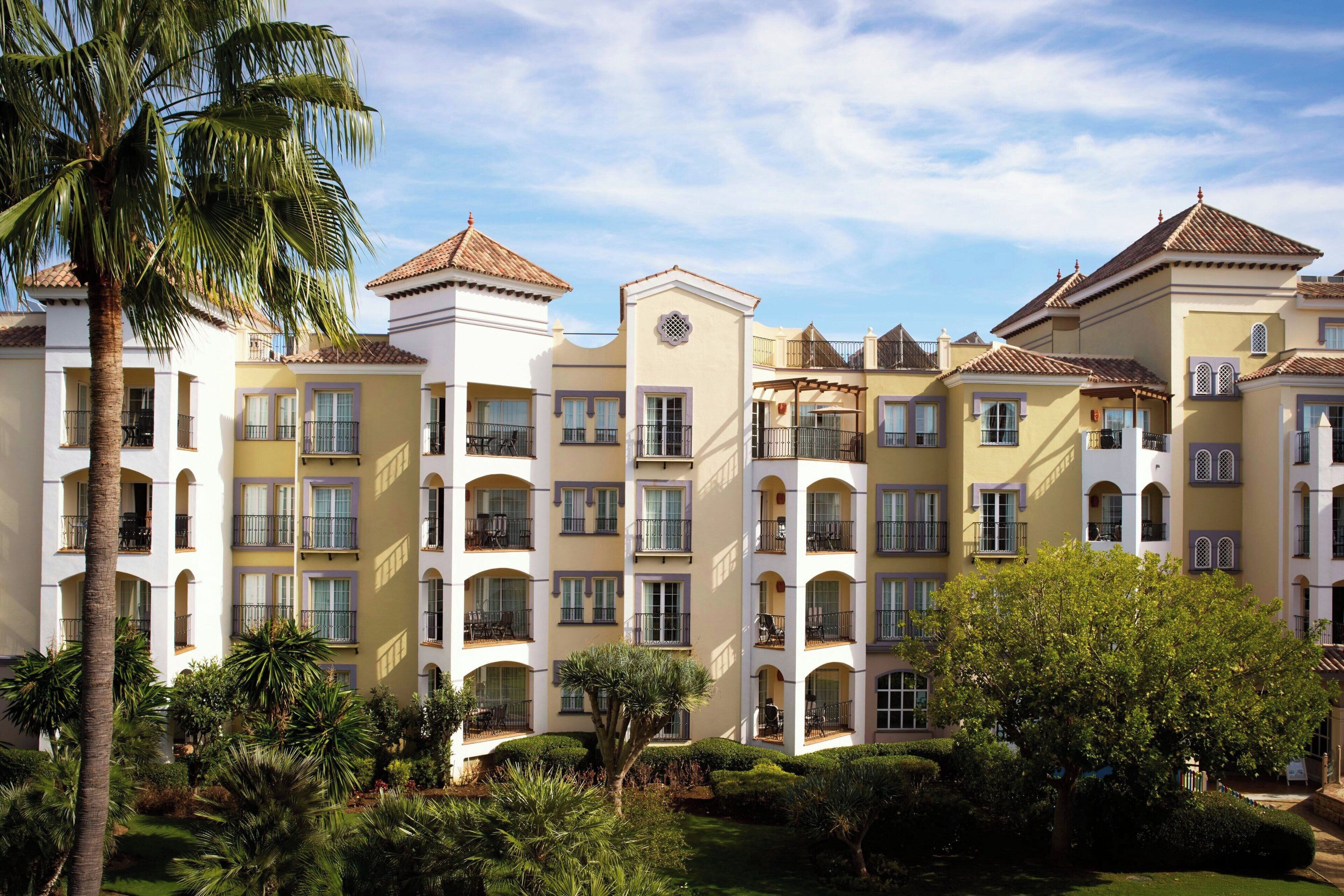 A multi-story hotel with balconies, a courtyard with palm trees, and a clear sky.