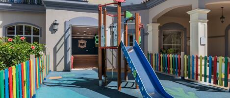 Zona de juegos infantiles con tobogán y estructura de escalada, rodeada de vallas de colores y la entrada de un edificio.