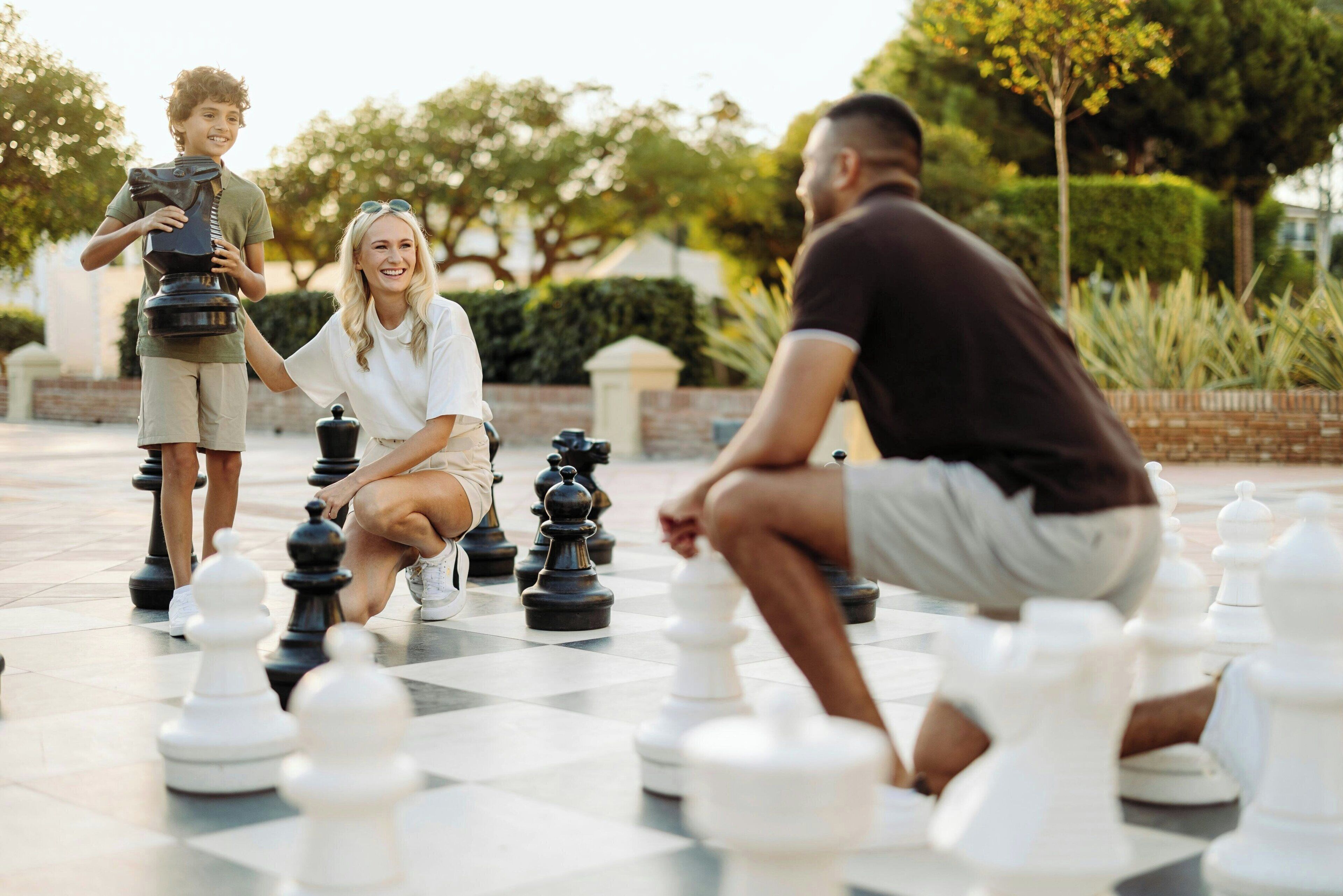 Three people playing chess outdoors on a checkered pavement.