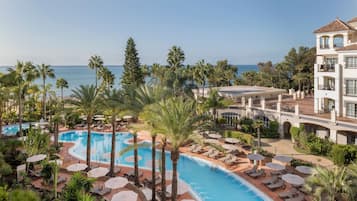 Et resort med en stor swimmingpool omgivet af palmer og liggestole, med udsigt over havet.