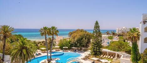 Piscina di un resort dalla forma sinuosa, circondata da palme e lettini, con vista sul mare e sugli edifici sullo sfondo.