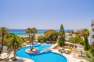 Piscina de diseño curvo en un resort, rodeada de palmeras y sillones de descanso, con vista al mar y edificios al fondo.