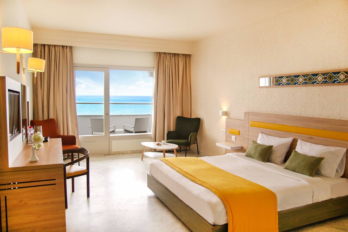 Een hotelkamer met een bed, een bureau, een stoel, een tafeltje en uitzicht op zee.