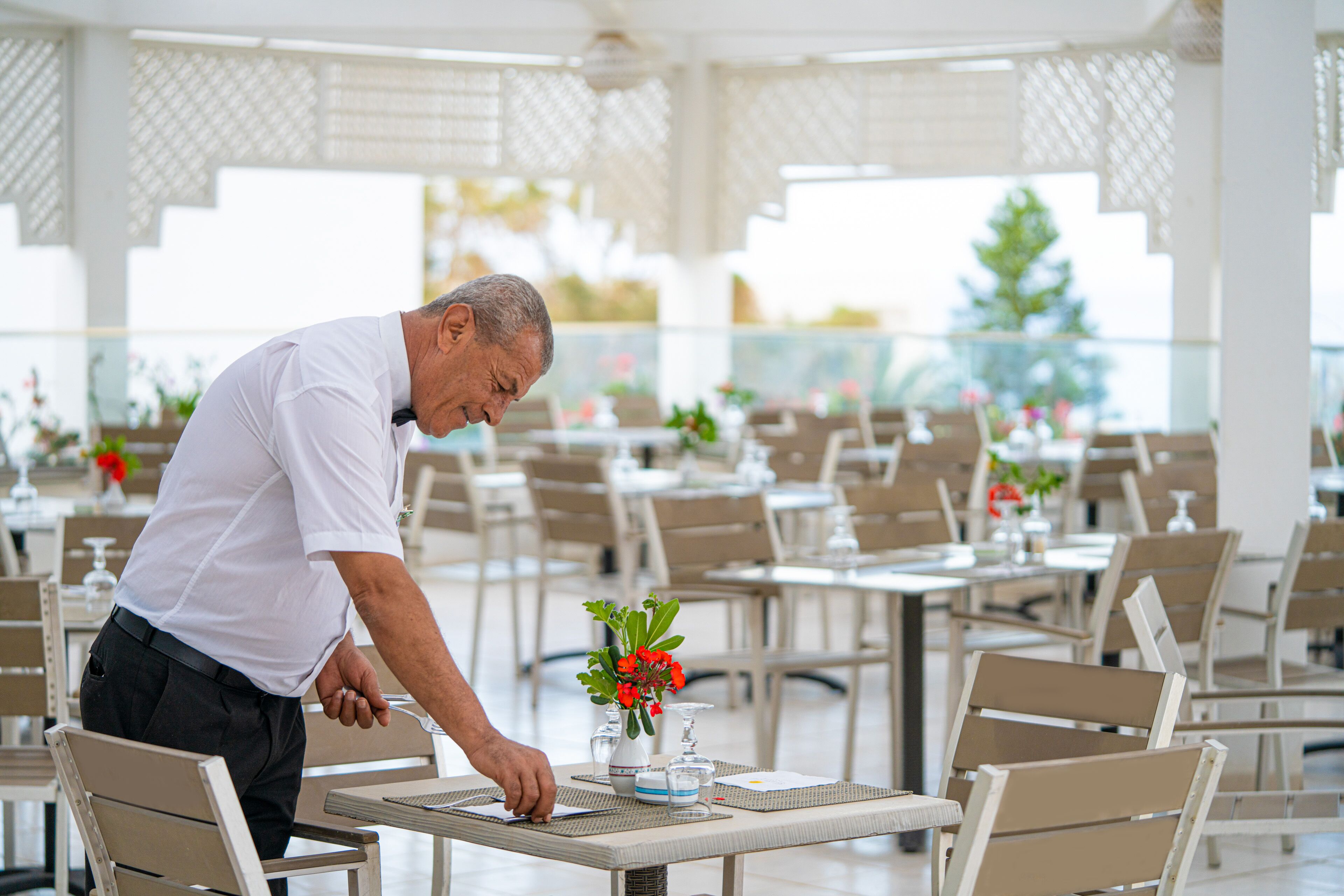 Ein Mann deckt einen Tisch in einem Restaurant ein, das mehrere Tische und Stühle bietet, mit einer dekorativen Vase und Blumen auf dem Tisch.