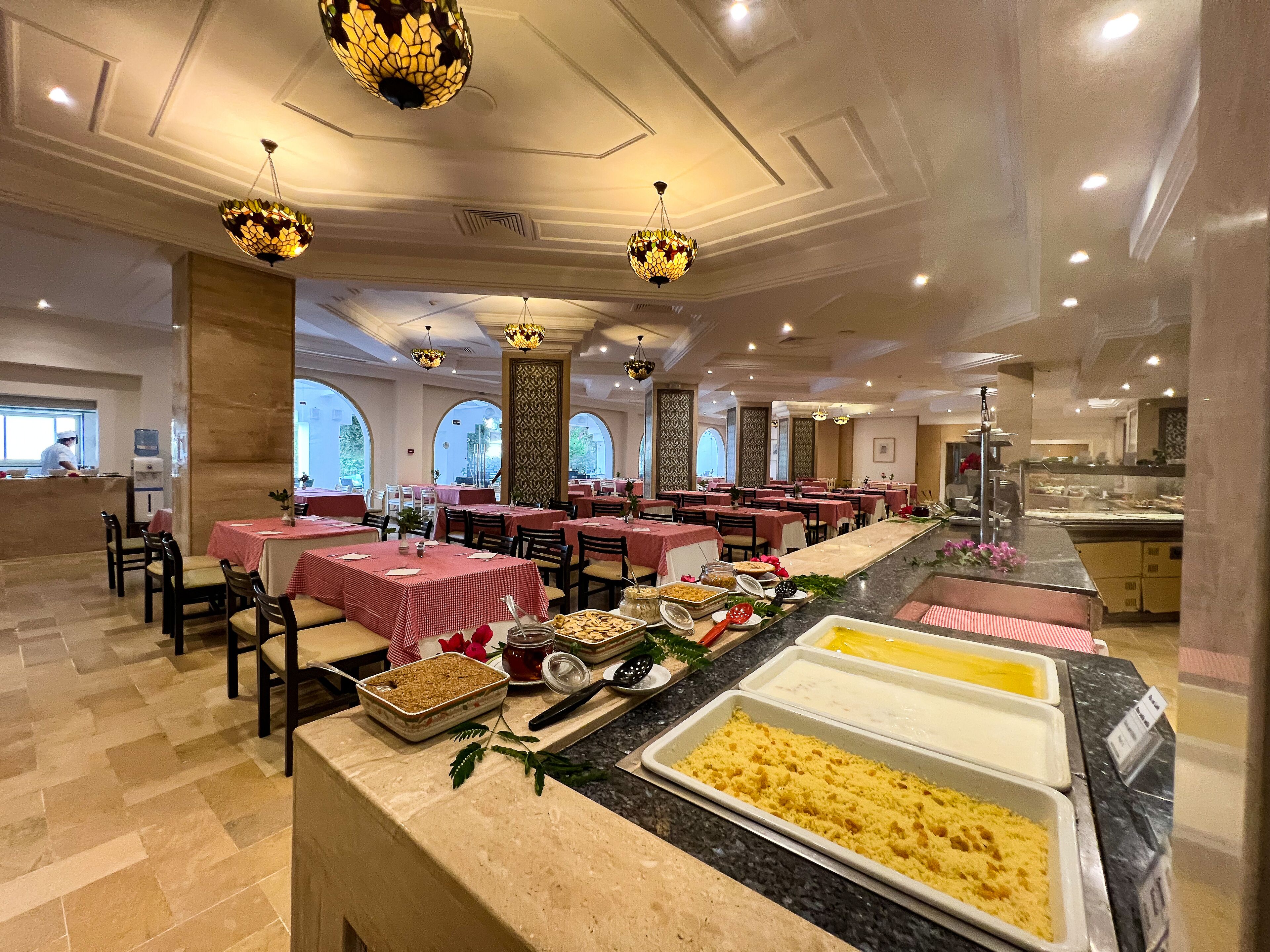 A spacious dining area with multiple tables, a buffet counter with various food items, and decorative lighting fixtures.