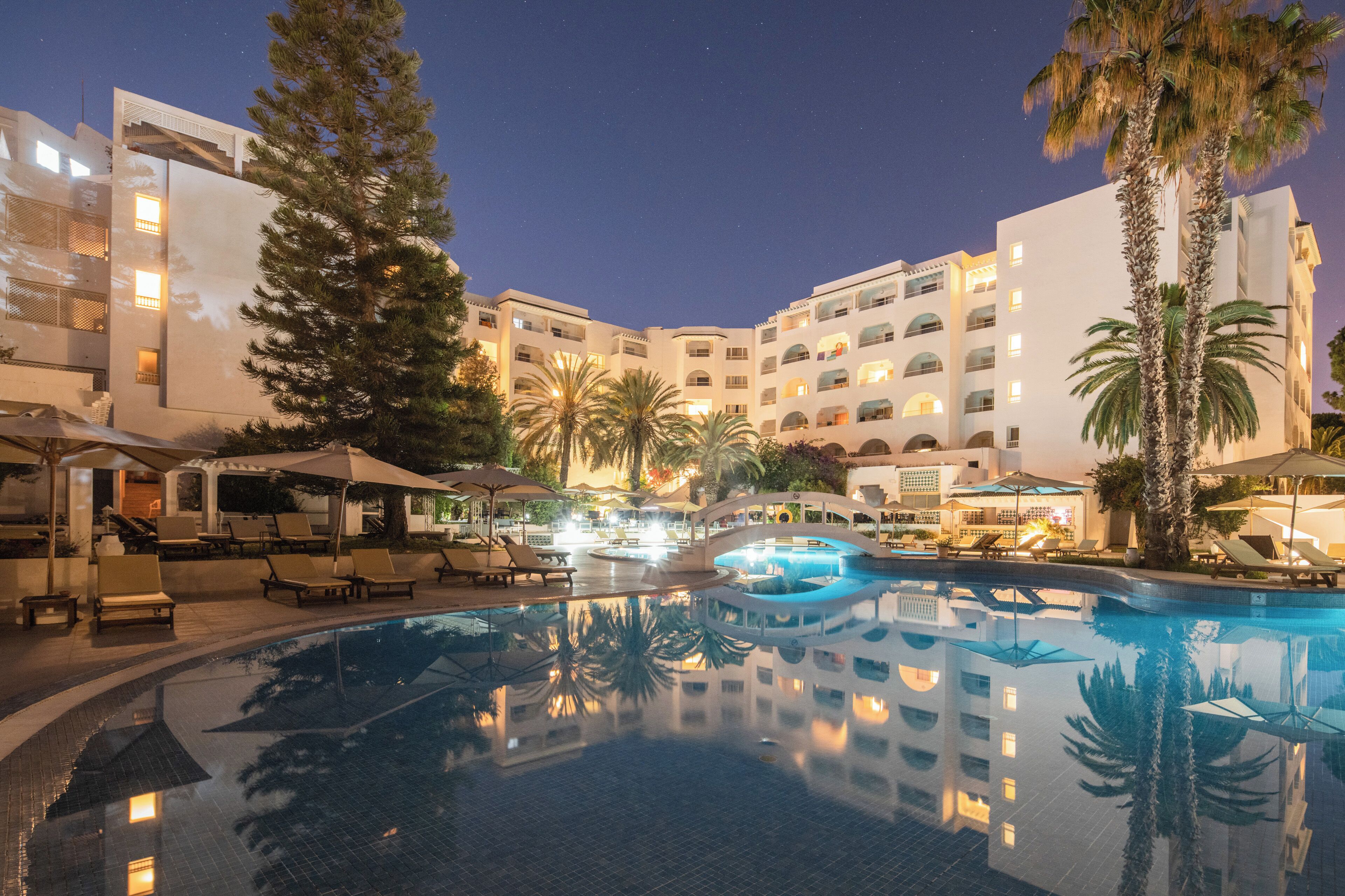 A hotel pool area with lounge chairs, umbrellas, and palm trees.