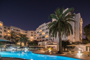 Vista nocturna de un área de piscina de hotel con sillas de descanso, sombrillas y una palmera.