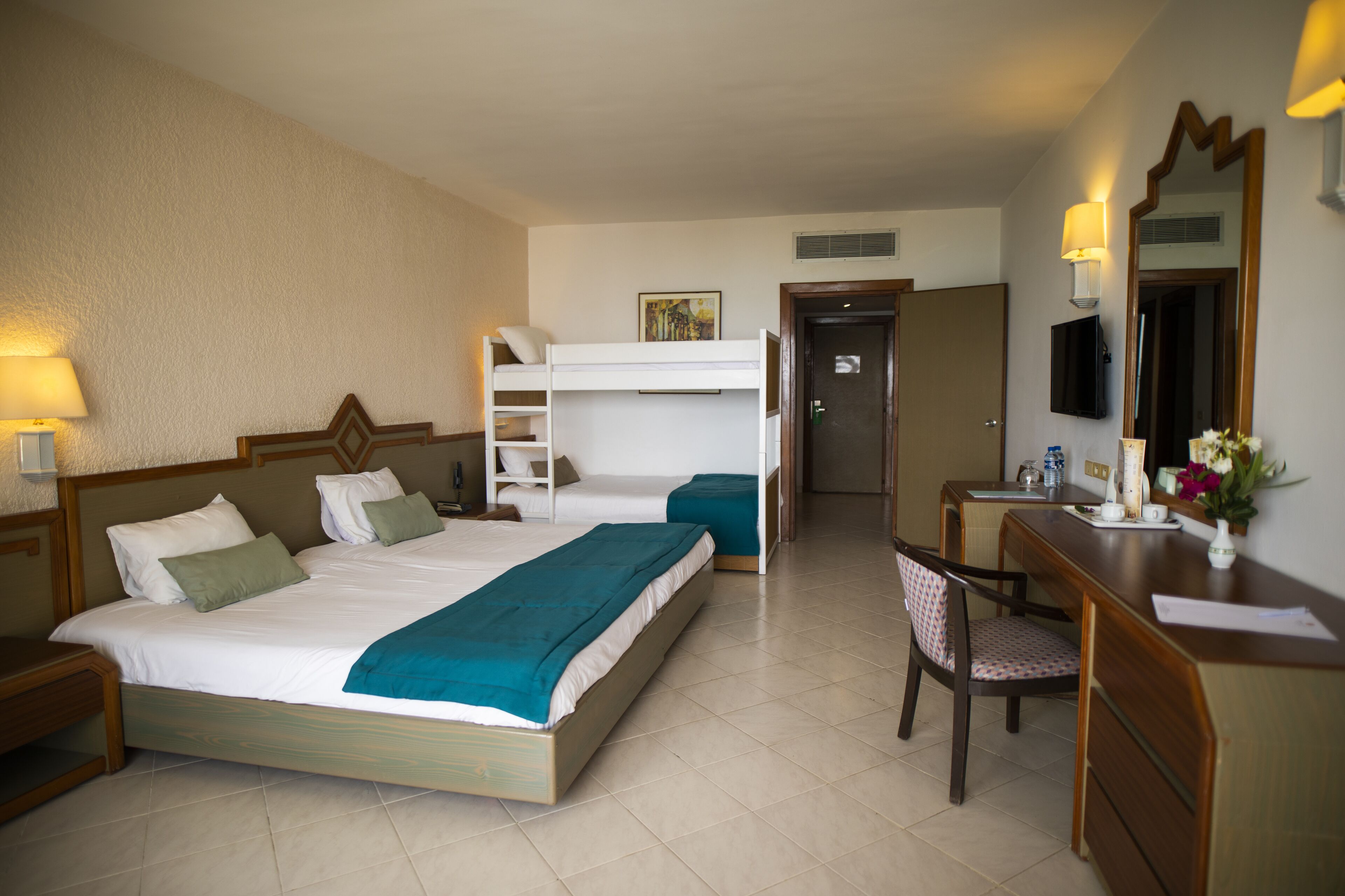 Habitación de hotel con cama, escritorio, silla, cómoda, espejo y puerta.