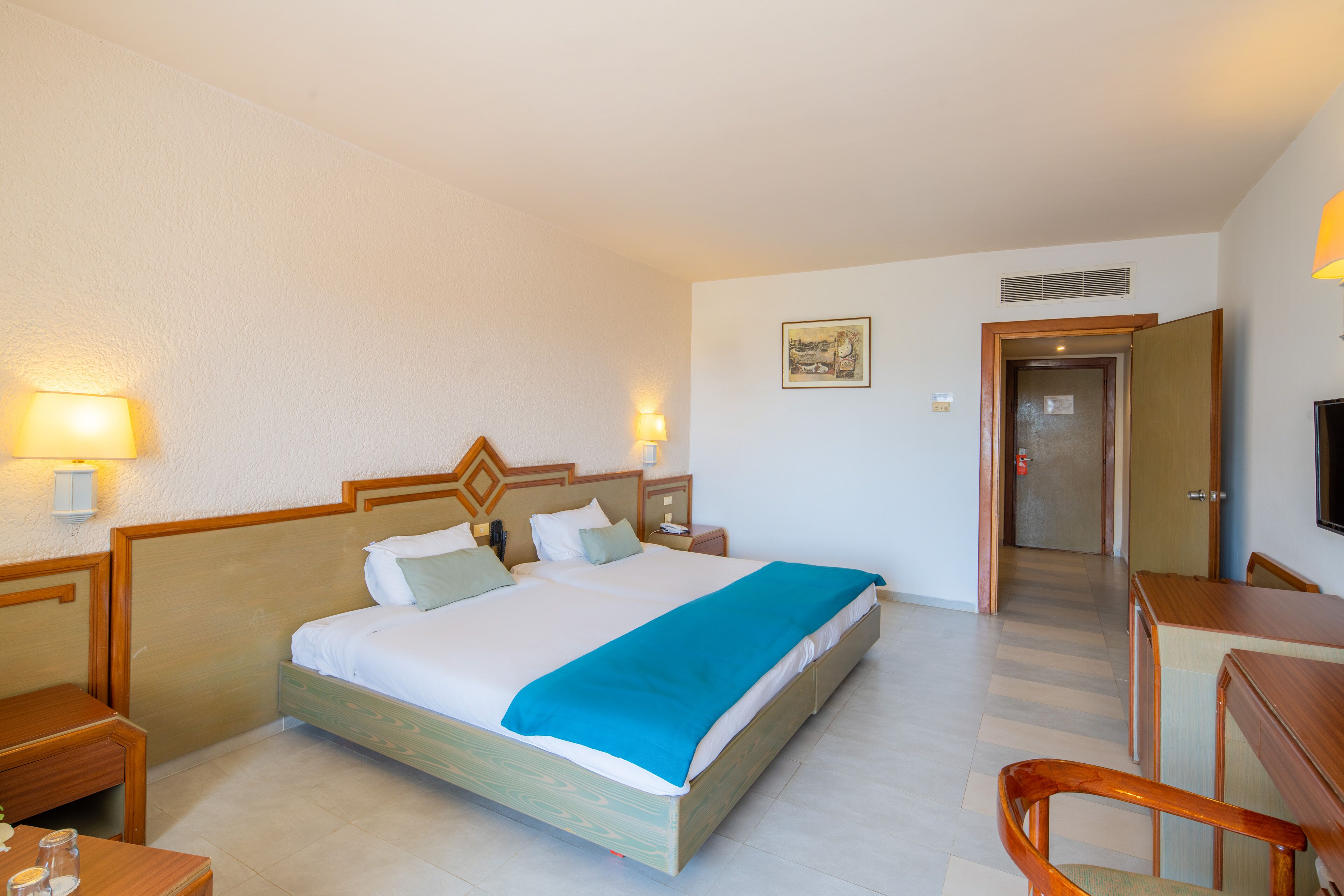 A hotel room with a large bed, bedside tables, a desk, and a chair. There is a door leading to another room and a wall-mounted TV.