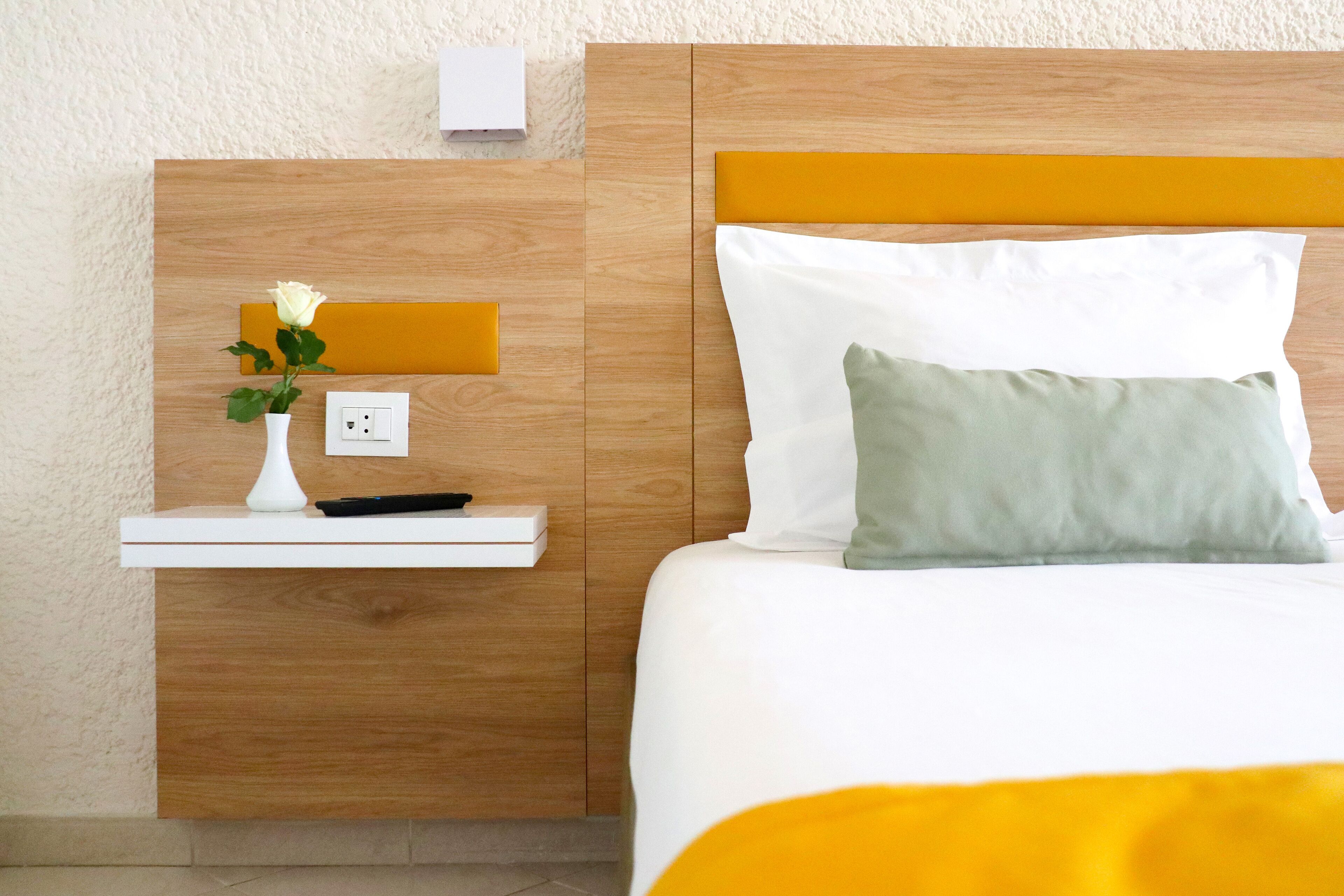 A neatly arranged hotel room with a wooden headboard, a bedside table with a vase of flowers, a remote control, and a power outlet.