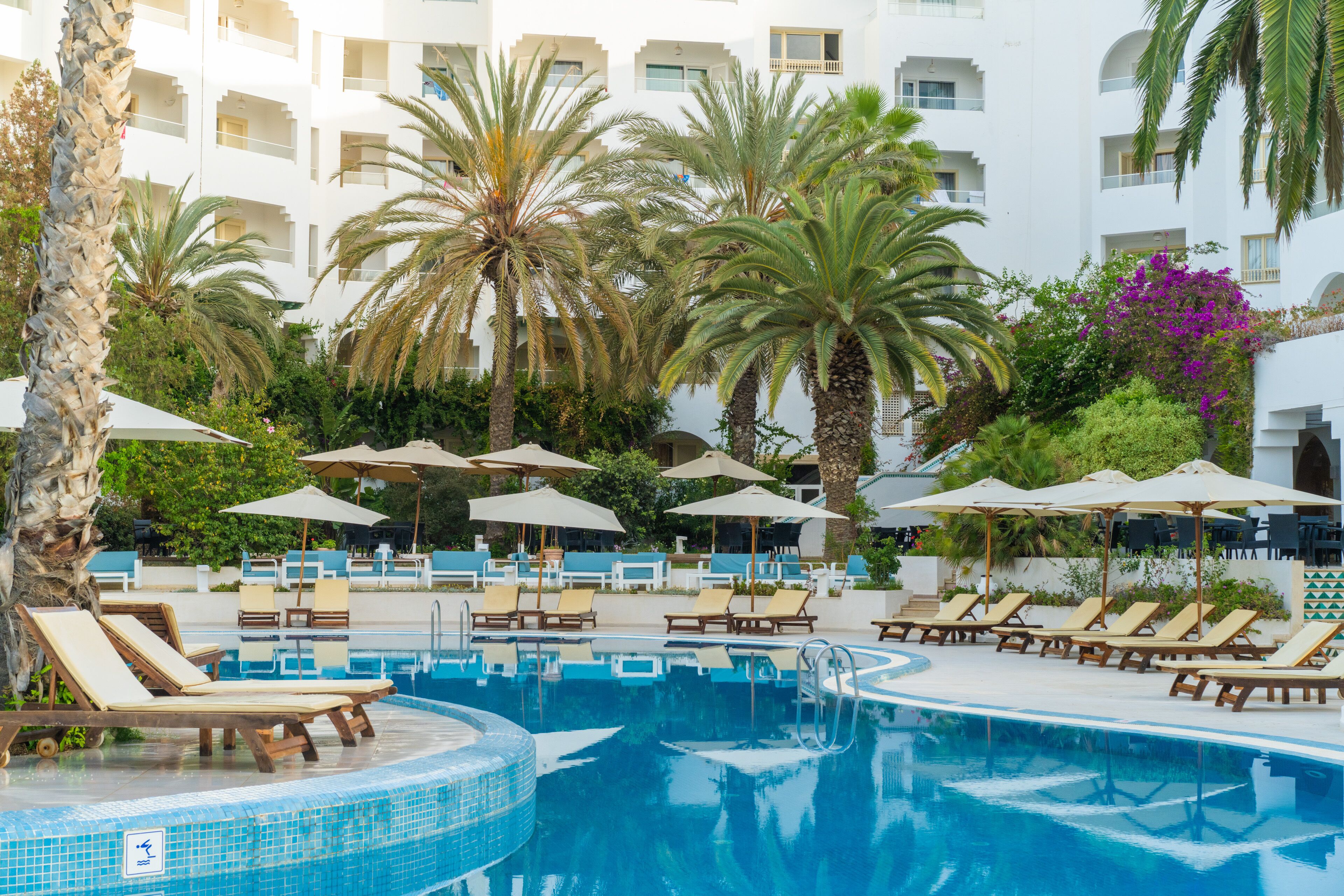 Un espace piscine agrémenté de chaises longues et de parasols, entouré de palmiers et bordé d’un bâtiment blanc doté de balcons.