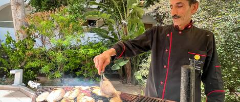 Un chef prépare différents types de viande sur un grand barbecue installé à l’extérieur, sous un auvent.