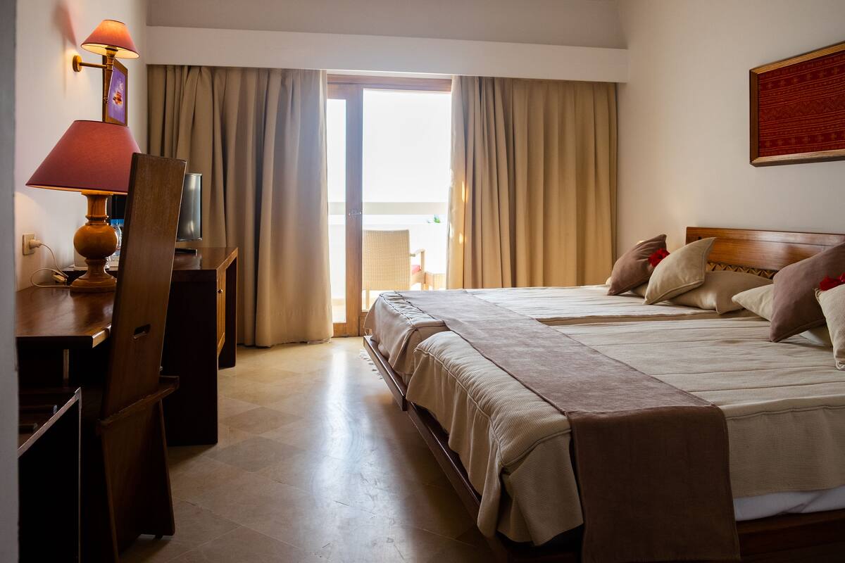 A hotel room with two beds, a desk with a lamp, a TV, and a view of the outdoors through a large window.