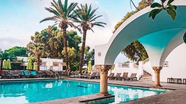 A swimming pool with lounge chairs and umbrellas, surrounded by palm trees and a white building with columns. - The Orangers Beach Resort and Bungalows - All Inclusive (Hammamet)