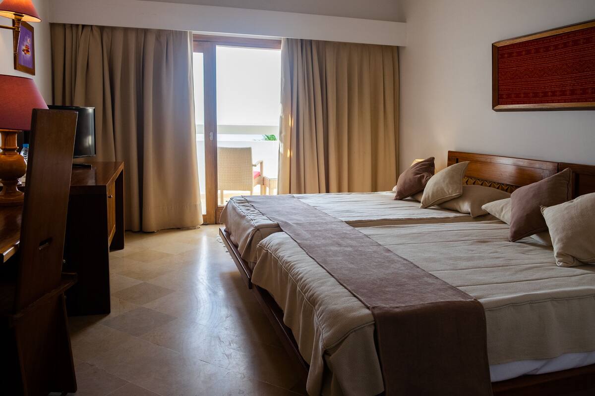 A hotel room with two beds, a desk with a lamp, and a view of the outdoors.